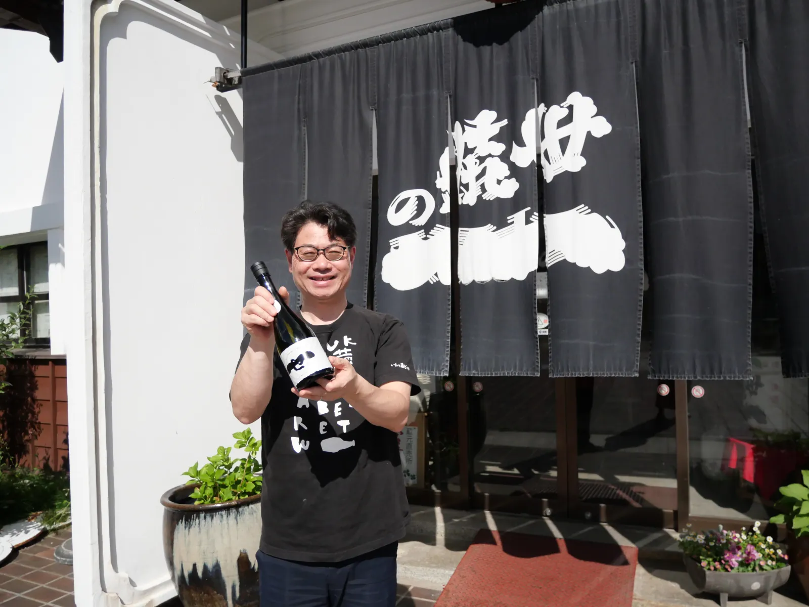 世嬉の一酒造の暖簾前で日本酒を手にする四代目社長 佐藤航